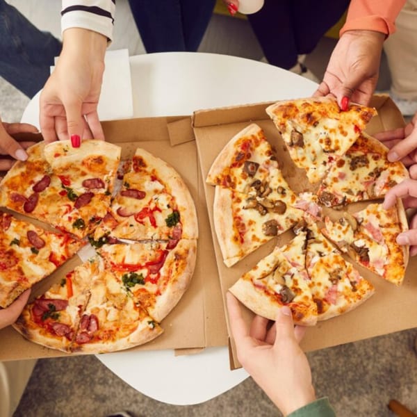 Multiple hands reaching for slices of two different pizzas in cardboard boxes on a white table.