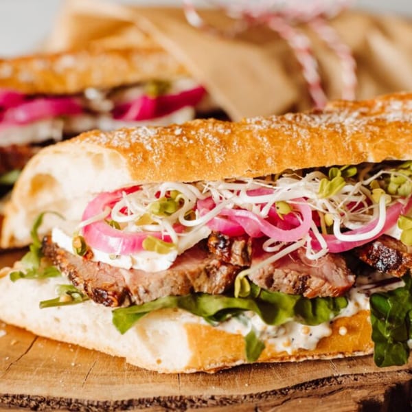 Close-up of a Zee's Kitchen steak sandwich with pickled onions, sprouts, and greens on a wooden board.