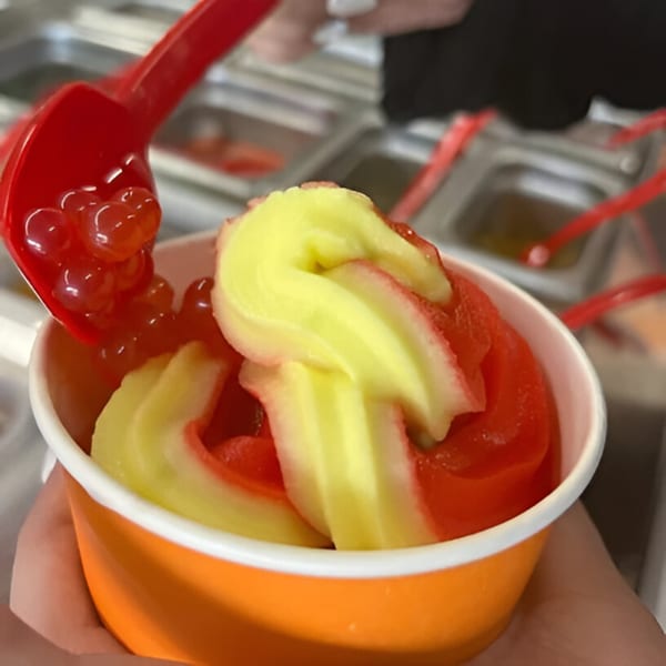 A person adding red boba pearls to a cup of swirled red and yellow frozen yogurt.