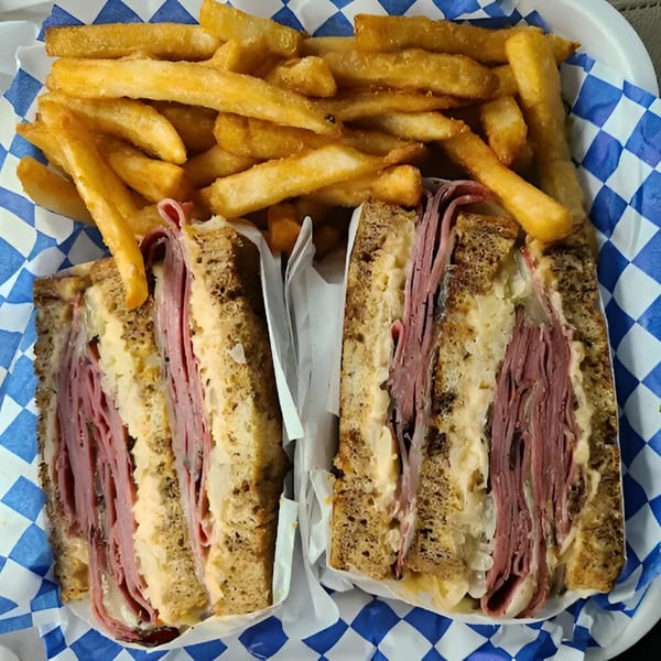 A Zee's Kitchen Reuben sandwich, cut in half, with a side of golden french fries in a blue and white checkered basket.