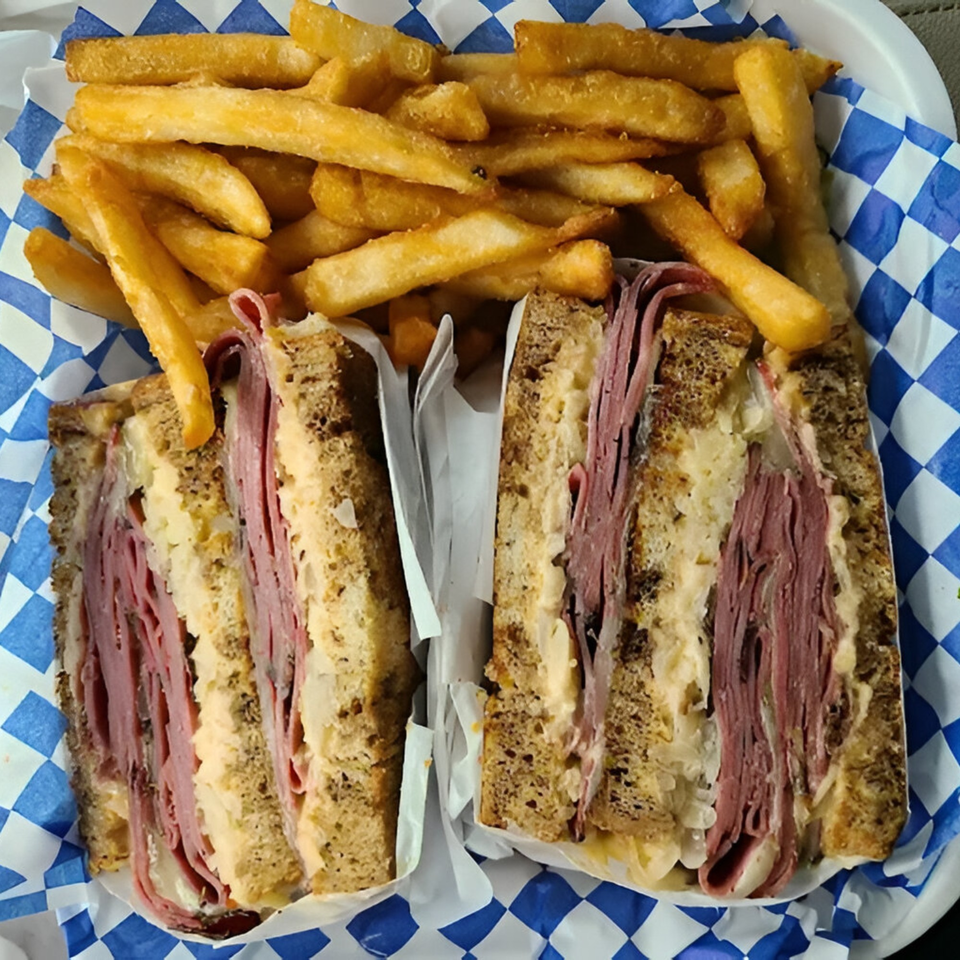 A Zee's Kitchen Reuben sandwich, cut in half, with a side of golden french fries in a blue and white checkered basket.